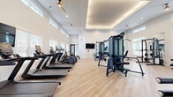 Located at 3100 Ascend Loop in Leland, NC, this fitness center offers a thoughtfully designed, single-floor layout comprising two dedicated exercise rooms. The facility is bright and airy, featuring soaring ceilings, clerestory windows, and abundant natural light streaming through large, shuttered windows that create an energizing atmosphere throughout.

The primary exercise room is generously proportioned, showcasing light hardwood-style flooring, a coffered tray ceiling with warm ambient lighting, and ceiling fans for added comfort. This space is fully equipped with a row of treadmills and elliptical machines along one wall, a multi-station cable machine, and a wall-mounted flat-screen TV. Sleek black-framed glass doors provide an elegant entry point into the space.

The secondary exercise room offers a complementary environment for strength and functional training. It features floor-to-ceiling mirrors, a ballet barre, and a curated selection of strength equipment including a Smith machine, adjustable benches, resistance machines, a dumbbell rack, exercise balls, and an Assault Fitness rowing machine. A built-in shelving unit with a mounted TV adds a refined, functional touch.

Together, these two rooms deliver a comprehensive, modern fitness experience suited for a wide variety of workout routines in a clean, well-appointed setting.