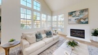 Located in Vista, CA, this two-story clubhouse offers a welcoming and well-appointed gathering space designed for comfort and functionality. The main floor hosts an open-concept layout that seamlessly connects a fully equipped kitchen, a dining area, and a spacious living room. The kitchen features white shaker cabinetry, quartz countertops, stainless steel appliances, and light hardwood flooring that flows throughout the space. The living room is a standout area, boasting soaring vaulted ceilings and a dramatic wall of floor-to-ceiling windows that flood the room with natural light and offer open views of the surrounding landscape. A fireplace adds a warm focal point to this bright, airy room. The dining area is thoughtfully furnished with a round wood table and cane-back chairs, complemented by a decorative sideboard and vibrant wall art. The upper floor provides additional flexible lounge space, complete with skylights, an open railing overlooking the lower level, and cozy seating areas adorned with tasteful wall décor. Throughout both floors, light hardwood flooring, recessed lighting, and a clean, neutral palette create a cohesive, modern ambiance ideal for community gatherings.