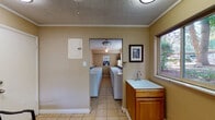 Located at 1216 Quivira Dr in Colorado Springs, CO, this single-floor shared laundry facility is designed for practicality and convenience. The space is organized into distinct functional zones, all finished with durable light-toned ceramic tile flooring and textured ceilings accented by crown molding throughout.

The main laundry room houses two parallel rows of commercial-grade top-loading washing machines and dryers, providing ample capacity for multiple users. Overhead ceiling fans and flush-mount light fixtures ensure the room remains well-lit and properly ventilated. Informational postings and laundry tips are displayed on the walls for user reference.

An adjoining utility area features a dedicated utility sink set into a warm wood-toned cabinet, alongside an electrical panel access point. A large window in this area overlooks mature trees and the surrounding outdoor landscape, bringing in natural light.

A separate waiting or transitional area provides seating with a small table and chair arrangement, a decorative wall niche, and additional windows that frame views of the tree-lined exterior. Baseboard heating units are present throughout, ensuring comfort during Colorado's cooler seasons. The overall layout is straightforward and efficient, making this facility a highly functional communal amenity.