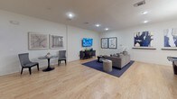 This generously sized game room offers a well-appointed environment designed for both entertainment and relaxation. Light hardwood flooring extends throughout the entire space, complemented by crisp white walls adorned with crown molding that adds a refined architectural touch. Recessed lighting illuminates the room evenly, enhancing its open, airy feel.

The room is organized into distinct zones. A central pool table serves as the focal point of the gaming area, accompanied by a wall-mounted cue rack nearby. Adjacent to the billiards area, a casual nook features high-top bar tables and industrial-style stools, ideal for spectators or social gatherings.

A dedicated lounge area anchors one end of the room, furnished with a plush gray sectional sofa, a navy geometric-patterned area rug, and a sleek dark credenza topped with decorative accents. A large flat-screen television is mounted above, making this corner well-suited for watching sports or films. A separate bistro table with accent chairs provides an intimate seating option. Curated wall art and decorative pieces, including an eye-catching circular wall installation, contribute to the space's sophisticated yet inviting ambiance.