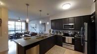 This contemporary single-floor residence showcases a thoughtfully designed open-concept layout that seamlessly connects the kitchen and living areas. The kitchen is a standout feature, boasting rich dark cabinetry that extends to the ceiling, offering generous storage space. Stainless steel appliances, including a range, microwave, and dishwasher, provide a polished, modern aesthetic. A large central island with a built-in sink and dark countertop serves as both a functional workspace and a casual dining area, accented by a trio of elegant pendant lights hanging overhead. Light hardwood flooring contrasts beautifully with the dark cabinetry, creating a warm yet sophisticated atmosphere. The open living area flows naturally from the kitchen, furnished with comfortable seating and anchored by tasteful wall art. Large windows allow natural light to filter into the space, enhancing the bright, airy ambiance. The overall design palette — combining deep espresso tones, cool stainless finishes, and neutral walls — creates a cohesive, upscale environment well-suited for both everyday living and entertaining.