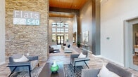 Located at 4001 Reedybrook Crossing in Apex, NC, the Flats 540 clubhouse is a thoughtfully designed community space that blends contemporary style with warm, inviting elements. The centerpiece of the space is a soaring two-story living area anchored by a striking stacked-stone column that extends from floor to ceiling, complemented by a warm wood-paneled ceiling, dramatic pendant lighting, and an abundance of natural light streaming through tall, multi-pane windows. Wide-plank hardwood-style flooring runs throughout, tying the open layout together cohesively. Multiple seating arrangements populate the main living area, offering both intimate and open gathering zones, while a built-in media wall with a mounted flat-screen TV provides a focal point for entertainment. The modern kitchen features dark cabinetry, stainless steel appliances, a large central island with bar seating, and stylish pendant lighting — ideal for social gatherings. A dedicated game room offers a relaxed retreat with plush seating, a wall-mounted TV, a large-format Scrabble board, and a pool table visible in the background. An additional media lounge area with a mounted TV and curated wall art rounds out the space. The overall ambiance is polished yet approachable, balancing upscale finishes with functional, community-oriented design.