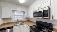 This single-story residence on British Square Drive in Winston-Salem, North Carolina, offers a well-organized layout with updated finishes throughout. The kitchen serves as a functional centerpiece, featuring white shaker-style cabinetry extending to the ceiling, white subway tile backsplash, and warm-toned countertops. Stainless steel appliances — including a refrigerator, electric range, over-the-range microwave, and dishwasher — provide a modern, cohesive look. A double-basin stainless steel sink sits beneath a window, allowing natural light into the space. Wood-look flooring runs through the kitchen and connects to an adjacent open area, which transitions into a spacious living room. A half-wall column separates the kitchen from the living space, creating definition while maintaining an open feel. The living room features rich dark hardwood-style flooring and windows that bring in natural light. Bedrooms are carpeted in neutral tones, each with windows and individual entry doors. Additional living areas display light wood-look flooring with clean gray walls throughout. The bathroom includes a white vanity with brushed nickel hardware, a full tub-shower combination with white tile surround, and wood-look flooring, completing the unit's consistent, refreshed aesthetic.