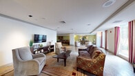 This expansive single-floor space is thoughtfully designed to accommodate a variety of social and recreational activities. The main lounge area serves as the heart of the facility, featuring warm hardwood flooring, recessed lighting with tray ceiling details, and multiple seating arrangements including plush sofas and accent chairs arranged around a central coffee table. A wall-mounted flat-screen television and a dark wood media console anchor one side of the room, while large windows dressed with decorative curtains allow natural light to fill the space.

The open floor plan flows seamlessly into a game room area, where a full-size billiards table takes center stage beneath warm ambient lighting. Adjacent to this is a cozy conversational seating nook with patterned upholstered chairs set against olive-toned accent walls. A kitchenette area is outfitted with dark wood cabinetry, granite countertops, stainless steel appliances, and pendant lighting overhead.

A dedicated media or theater room features plush sectional seating, patterned carpeting, and cinema-themed wall décor, creating an inviting screening environment. Additional transitional corridors with carpeted flooring and sliding doors provide flexible room separation throughout the facility.