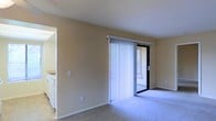 This single-floor residence offers a clean, functional layout designed for comfortable everyday living. The main living area is carpeted and bathed in natural light, thanks to a full-height sliding glass door fitted with vertical blinds that open to an outdoor space, providing a seamless connection between interior and exterior. The neutral beige walls create a calm, welcoming ambiance throughout the room, while the light-colored carpet adds warmth underfoot. Adjacent to the living area, the kitchen is visible through an open passageway, revealing white cabinetry and countertops that contribute to a bright, orderly feel. The kitchen's layout appears practical and efficient, with ample cabinet storage. The overall design of the space is straightforward and uncluttered, with smooth walls and simple finishes that lend themselves well to personalization. The abundant natural light filtering through the sliding door enhances the sense of openness throughout the living area, making the space feel airy and inviting. The single-floor layout ensures ease of navigation, offering a cohesive flow between the living and kitchen areas.