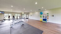 Located at 36 Broadway Village Dr in Columbia, Missouri, this fitness center offers a well-equipped, single-floor exercise space designed to support a wide range of workout routines. The open layout is finished with warm wood-look flooring throughout, complemented by protective rubber mat zones strategically placed beneath equipment for safety and durability.

Large windows along multiple walls allow abundant natural light to fill the space and provide views of the surrounding greenery, creating an energizing atmosphere. Recessed ceiling lighting ensures even illumination throughout the room.

The cardio section is well-appointed, featuring treadmills, elliptical trainers, a stationary bike, and rowing machines. A wall-mounted flat-screen television is positioned for convenient viewing during workouts. Strength training needs are addressed with a cable machine, a weight bench, and a full rack of dumbbells neatly organized along a mirrored wall, which also helps to visually expand the space.

A dedicated flexibility and functional fitness area includes a rack stocked with stability balls in multiple sizes, resistance bands, foam rollers, and medicine balls. A wall-mounted hook rack provides convenient storage for personal items. Glass entry doors offer easy access, and posted fitness center rules help maintain an orderly environment. Overall, the space is clean, modern, and thoughtfully arranged to accommodate diverse fitness needs.