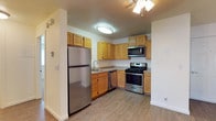 This 2-bedroom, 2-bathroom model unit is thoughtfully designed to offer comfortable, functional living across a single floor. The kitchen serves as a highlight of the space, featuring a full suite of stainless steel appliances including a gas range, over-the-range microwave, dishwasher, and a full-size refrigerator. Warm-toned wood cabinetry lines the walls, providing generous storage both above and below the counter, while the light-colored countertops offer ample prep space. A stainless steel sink is positioned beneath a dedicated light fixture, and a ceiling fan with integrated lighting overhead adds comfort and airflow to the area. The kitchen transitions seamlessly into the adjacent living areas, thanks to an open layout framed by clean white walls and neutral hardwood-style flooring that runs throughout the unit, lending a cohesive and modern feel. Textured ceilings add a classic touch to the interior. The overall design balances practicality with a welcoming aesthetic, making this model unit a strong representation of the comfortable, move-in-ready living experience available in this community.