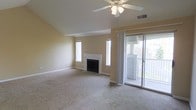 This single-floor space centers around a spacious living area characterized by vaulted ceilings that lend a sense of openness and volume. A white-mantled fireplace serves as the room's focal point, flanked by two vertically oriented windows that draw in natural light and contribute to the bright, airy feel of the interior. The living area is finished with wall-to-wall carpet in a neutral tone, complementing the warm beige walls throughout.

A ceiling fan with an integrated light fixture is centrally positioned, providing both airflow and ambient lighting. The room benefits from multiple light sources, including natural light streaming through the windows and the sliding glass door. This door opens directly onto a balcony, offering outdoor space with a railing enclosure and views of the surrounding exterior.

The sliding glass door is framed by a sheer curtain panel, adding a soft, functional touch to the transition between indoor and outdoor spaces. Overall, the living area presents a clean, neutral palette with practical features, making it a versatile and comfortable environment.