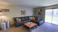 This single-floor residence offers a comfortable and thoughtfully styled living space with a distinct personality. The living room serves as the centerpiece of the home, featuring a large dark leather sectional sofa accented with orange and patterned throw pillows that add a pop of color and warmth. An industrial-style coffee table on caster wheels, painted in a bold orange finish, serves as a unique focal point and reinforces the eclectic aesthetic of the room. The carpeted flooring adds softness underfoot, contributing to the cozy feel of the space. Wall décor includes a creative collage-style clock display and an abstract canvas print, lending an artistic touch to the neutral-toned walls. A turquoise shelving unit in the corner provides both storage and a contrasting accent color, complementing the orange accents throughout. Natural light filters in through a sliding glass door or large window along one wall, brightening the room and offering a connection to the outdoors. The overall ambiance of this residence is casual yet curated, blending comfort with creative design choices that make the space feel lived-in and inviting.