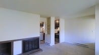 This single-floor residence offers a comfortable and functional layout that blends the living and kitchen areas in an open, airy arrangement. The living room is anchored by a classic brick fireplace with a dark metal screen, adding warmth and a traditional focal point to the space. Neutral-toned carpeting covers the living area floor, contributing to a calm and inviting atmosphere throughout.

A built-in shelving unit serves as a partial divider between the living room and the kitchen, providing both practical storage and a visual transition between the two areas. The kitchen features dark cabinetry that contrasts cleanly with light-colored walls, and is equipped with stainless steel appliances, including a refrigerator and a black range. The kitchen floor transitions to hardwood-style flooring, offering a subtle distinction from the carpeted living space.

Baseboard heating units are present along the walls, ensuring efficient climate control throughout the home. The overall color palette — soft whites, warm neutrals, and dark wood tones — creates a cohesive and understated aesthetic. The space is well-suited for everyday living, with a straightforward design that emphasizes usability and a welcoming ambiance.
