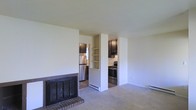 This single-floor residence offers a comfortable and functional layout that blends the living and kitchen areas in an open, airy arrangement. The living room is anchored by a classic brick fireplace with a dark metal screen, adding warmth and a traditional focal point to the space. Neutral-toned carpeting covers the living area floor, contributing to a calm and inviting atmosphere throughout.

A built-in shelving unit serves as a partial divider between the living room and the kitchen, providing both practical storage and a visual transition between the two areas. The kitchen features dark cabinetry that contrasts cleanly with light-colored walls, and is equipped with stainless steel appliances, including a refrigerator and a black range. The kitchen floor transitions to hardwood-style flooring, offering a subtle distinction from the carpeted living space.

Baseboard heating units are present along the walls, ensuring efficient climate control throughout the home. The overall color palette — soft whites, warm neutrals, and dark wood tones — creates a cohesive and understated aesthetic. The space is well-suited for everyday living, with a straightforward design that emphasizes usability and a welcoming ambiance.