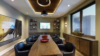 This single-floor space showcases a thoughtfully designed interior centered around a standout conference or dining room. The focal point is a substantial live-edge wood table, its natural grain and organic form lending warmth and character to the space. Surrounding it are sleek black leather chairs that offer both comfort and a contemporary aesthetic.

Overhead, a geometric coffered ceiling with warm wood paneling and integrated LED accent lighting creates a dramatic architectural statement. A cylindrical pendant light hangs centrally, adding to the layered lighting design alongside recessed ceiling fixtures.

The walls are lined with floating shelves displaying decorative ceramic vases in muted and bold tones, adding curated visual interest. A wall-mounted flat-screen TV is centrally positioned, making the room well-suited for presentations or media viewing. An exposed brick accent wall introduces texture and an industrial contrast to the otherwise polished interior.

Natural light filters in through a window on one side, while rustic wood cabinetry beneath provides functional storage. The combination of natural materials, warm lighting, and refined furnishings gives the space a professional yet inviting ambiance.