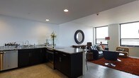 This single-floor space showcases a sleek, modern design centered around an open-concept layout that seamlessly connects the kitchen and living area. The kitchen is a standout feature, boasting dark espresso cabinetry, polished granite countertops with subtle veining, and a central island that doubles as a casual dining and prep space. Recessed lighting in the smooth white ceiling keeps the kitchen bright and functional. Stainless steel appliances add a contemporary touch throughout.

The living area flows naturally from the kitchen, featuring polished concrete-style flooring accented by a rich, deep-red area rug that adds warmth to the otherwise clean, neutral palette. A tufted charcoal sofa and complementary armchairs create an inviting seating arrangement, anchored by a decorative round mirror with a bold dark frame that draws the eye along the wall.

Floor-to-ceiling windows along the far wall flood the living space with abundant natural light and offer expansive outward views, enhancing the sense of openness and airiness throughout. The minimalist white walls provide a crisp backdrop that allows the dark furnishings and architectural details to stand out. Overall, the space strikes a sophisticated balance between urban modern aesthetics and comfortable, livable design.