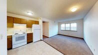 This single-floor space offers a functional and straightforward layout that combines the kitchen and living area in an open, connected fashion. The kitchen is equipped with warm honey-toned wood cabinetry providing ample storage, white appliances including a refrigerator and electric range, and a ventilation hood above the stove. The flooring in the kitchen area is composed of light-toned tile, offering easy maintenance and a clean aesthetic. Transitioning seamlessly from the kitchen, the living area features wall-to-wall carpeting that lends a sense of warmth and comfort underfoot. A multi-pane window along the far wall allows natural light to fill the living space, creating a bright and welcoming atmosphere. The ceilings feature a subtle textured finish throughout, and flush-mount light fixtures provide consistent, even illumination. A gently arched passageway connects the main living area to an adjoining space, adding a soft architectural detail to the otherwise simple design. The overall ambiance is practical and comfortable, with a neutral color palette of whites and beiges that provides a versatile backdrop suitable for a variety of furnishing styles.