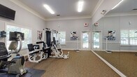 Located at 1878 Gingercake Cir in Rock Hill, SC, this single-floor fitness center is thoughtfully designed to support a well-rounded workout experience. The space features neutral gray walls accented by crisp white crown molding and chair rail detailing, lending the room a polished, welcoming feel. Carpeted flooring runs throughout, providing cushioning underfoot during exercise sessions.

The fitness area is equipped with a multi-station strength training machine, allowing users to perform a variety of resistance exercises in one central location. A dedicated cardio zone houses an elliptical trainer, a treadmill, and an upright stationary bike, each with individual digital display consoles. A wall-mounted flat-screen television is positioned to keep users entertained during cardio workouts.

Large windows fitted with blinds line multiple walls, flooding the space with natural light and offering views of the surrounding greenery. A full-length wall mirror on one side of the room enhances the sense of spaciousness while allowing users to monitor their form. Fitness-themed artwork is displayed throughout, adding motivational energy to the environment.

Access to an adjacent common area is provided through elegant white French doors with glass panels, while a secondary exterior door offers convenient direct outdoor access. The overall ambiance is bright, clean, and purpose-built for fitness.