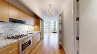 Located at 95 Wall Street, Unit 2120 in the heart of Lower Manhattan, this 390-square-foot studio apartment offers a well-designed single-floor layout comprising a living area, bathroom, and closet. The open-plan living space features warm honey-toned hardwood floors throughout, crisp white walls, and large floor-to-ceiling windows that flood the room with natural light and frame striking views of the surrounding Manhattan skyline. The integrated kitchen is outfitted with flat-front natural oak cabinetry, marble countertops and a matching marble backsplash, stainless steel appliances including a gas range, built-in microwave, and refrigerator — all accented by an elegant crystal chandelier overhead. The bathroom is a standout feature, clad entirely in veined white marble across the floors and walls, and appointed with a floating oak vanity, a modern vessel sink, and a lit mirror, creating a luxurious spa-like atmosphere. A dedicated closet provides practical storage within the compact footprint. The unit's elevated position delivers panoramic city views, making this a sophisticated urban retreat in one of New York City's most iconic financial district addresses.