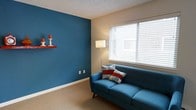 This single-floor space offers a compact yet well-designed living area that blends modern style with comfortable functionality. The living room serves as the focal point of the space, distinguished by a striking deep teal accent wall that adds a bold splash of color and visual depth. A decorative floating shelf mounted on the accent wall provides a tasteful display surface, currently adorned with a set of red candle holders that complement the room's color palette. The remaining walls are finished in a neutral beige tone, creating a balanced contrast that keeps the space feeling open and inviting. Soft, wall-to-wall carpeting in a warm tan hue covers the floor, contributing to the room's cozy ambiance. A large window fitted with white horizontal blinds allows for abundant natural light to filter in, brightening the interior throughout the day while offering adjustable privacy. The textured popcorn ceiling adds a classic architectural detail overhead. Overall, the space presents a cheerful and well-coordinated interior with a contemporary aesthetic, making it a comfortable and practical living environment.