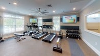 Located at 9001 High Peak Dr. in Greenville, SC, this fitness center is a comprehensively outfitted facility designed to support a wide range of workout routines. The space is organized into distinct training zones, all finished with durable rubber-blend flooring and united by a clean, neutral color palette with crisp crown molding and white trim throughout.

The cardio zone features a row of True-brand treadmills and elliptical machines, each positioned in front of large windows fitted with plantation shutters that invite natural light and views of the surrounding greenery. Wall-mounted flat-screen televisions provide entertainment during workouts. Ceiling fans contribute to air circulation and comfort throughout the space.

The strength training area includes a multi-function cable machine and an adjustable weight bench, while a full dumbbell rack offers a comprehensive selection of free weights. A large wall-mounted mirror allows users to monitor their form. A dedicated functional training section houses TRX suspension trainers, resistance bands, medicine balls, stability balls, and kettlebells neatly organized on a wall-mounted rack.

Recessed lighting supplements the abundant natural light, creating a bright and energizing atmosphere. The facility also includes a mirrored wall feature and direct access to an adjacent room, enhancing the overall flow and usability of the space.