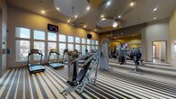 This fitness center offers a comprehensive workout environment designed to accommodate a wide range of exercise routines. The main floor is generously sized, featuring soaring ceilings with decorative circular ceiling details, ceiling fans, and recessed lighting that create an open, energizing atmosphere. Bold accent walls in warm orange and yellow tones add vibrancy to the space, while floor-to-ceiling windows along multiple walls flood the interior with natural light and offer outdoor views of the surrounding landscape. The distinctive striped carpeting throughout ties the space together visually.

The main workout area is outfitted with a full lineup of Cybex treadmills, ellipticals, and recumbent bikes, complemented by Hoist cable machines and multi-function strength stations. A dedicated free weight section includes a tiered dumbbell rack, weight bench, and barbell equipment, all positioned beside mirrored walls to support proper form. Wall-mounted televisions are strategically placed throughout for entertainment during workouts.

A separate glass-enclosed studio features spin bikes, large windows, and a ceiling fan, and is designated as a yoga lounge — ideal for group classes or individual training. Additional amenities include medicine balls, stability balls, and a stretching area, making this fitness center a thorough and well-rounded facility.
