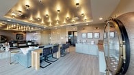 This elegantly designed single-floor space combines contemporary style with versatile functionality across an open, cohesive layout. The most striking architectural feature is the expansive tray ceiling adorned with an array of globe pendant lights that cast a warm, ambient glow throughout the main area, creating a distinctly upscale atmosphere.

The main open area integrates a comfortable lounge zone anchored by a plush sectional sofa in muted blue-gray tones, paired with a sleek gold accent table. Adjacent to the lounge sits a long communal dining table with modern cantilever chairs, ideal for group gatherings. A wet bar with sage-green cabinetry and a built-in sink provides convenient service functionality nearby.

A semi-private nook is separated from the main space by a textured, translucent room divider, housing a round wooden table with upholstered chairs and vintage-style framed artwork, lending a more intimate setting for smaller meetings or casual use.

A wall of floor-to-ceiling black-framed glass doors opens to a spacious balcony, flooding the interior with natural light and offering elevated outdoor views of the surrounding tree-lined landscape. Neutral-toned carpet and warm wood accents throughout unify the design, while decorative elements—including a circular mirrored display cabinet and curated wall art—add personality and visual interest.