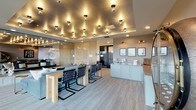 This elegantly designed single-floor space combines contemporary style with versatile functionality across an open, cohesive layout. The most striking architectural feature is the expansive tray ceiling adorned with an array of globe pendant lights that cast a warm, ambient glow throughout the main area, creating a distinctly upscale atmosphere.

The main open area integrates a comfortable lounge zone anchored by a plush sectional sofa in muted blue-gray tones, paired with a sleek gold accent table. Adjacent to the lounge sits a long communal dining table with modern cantilever chairs, ideal for group gatherings. A wet bar with sage-green cabinetry and a built-in sink provides convenient service functionality nearby.

A semi-private nook is separated from the main space by a textured, translucent room divider, housing a round wooden table with upholstered chairs and vintage-style framed artwork, lending a more intimate setting for smaller meetings or casual use.

A wall of floor-to-ceiling black-framed glass doors opens to a spacious balcony, flooding the interior with natural light and offering elevated outdoor views of the surrounding tree-lined landscape. Neutral-toned carpet and warm wood accents throughout unify the design, while decorative elements—including a circular mirrored display cabinet and curated wall art—add personality and visual interest.