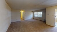 This single-floor residence presents a straightforward, functional layout spread across several interconnected rooms. The living area is generously proportioned, featuring wall-to-wall carpeting in a neutral beige tone and smooth, light-colored walls that create a clean, open atmosphere. A large window allows natural light to filter into the space, offering views of surrounding greenery. The floor plan, visible in the dollhouse overview, reveals a logical flow connecting the living area, bedrooms, kitchen, and bathrooms.

The bedrooms are carpeted and similarly finished in neutral tones, each featuring windows fitted with horizontal blinds. Closet access is provided, and the rooms offer adequate space for standard furnishings. The kitchen is equipped with warm oak cabinetry, dark laminate countertops, a stainless steel sink, a white electric range, and a dishwasher. Tile flooring in the kitchen provides a practical contrast to the carpeted areas elsewhere.

Two bathrooms serve the unit. One features a white vanity with under-cabinet storage, a mirrored medicine cabinet with overhead lighting, and tile flooring. The second bathroom includes a full bathtub with tiled surround, a toilet, and a vanity with cabinet storage. Both bathrooms are finished in light, neutral tones, contributing to the overall clean and understated character of the space.