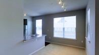 This single-floor space showcases an open-concept design that seamlessly connects the kitchen and living areas. The kitchen features a peninsula with granite countertops and an integrated sink, providing both workspace and a casual dining option. Stainless steel appliances, including a full-size refrigerator, add a modern, functional touch, while a dedicated door likely leads to additional storage or utility space.

Track lighting runs along the ceiling, offering adjustable illumination throughout the kitchen and living areas. The living space is finished with neutral wall-to-wall carpeting that contributes to a warm, comfortable feel. Two generously sized windows fitted with white horizontal blinds allow abundant natural light to fill the room while maintaining privacy. The grey-toned walls create a contemporary, neutral backdrop that complements the overall design.

The open layout promotes a sense of spaciousness and ease of movement between the kitchen and living areas, making the space practical for everyday living and entertaining.