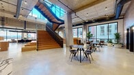 This thoughtfully designed commercial space blends industrial and contemporary aesthetics across a single, expansive floor. The architecture is defined by exposed concrete ceilings, structural columns, and polished concrete floors that establish a raw yet refined foundation throughout. Overhead, open ductwork and track lighting reinforce the industrial character while ensuring the space remains well-lit and functional.

A striking centerpiece staircase constructed from warm wood treads and bold steel framing rises dramatically beneath a skylight, flooding the surrounding open area with natural light. Adjacent to the staircase, a tiered wood platform with cushioned seating provides a casual gathering or lounge area. Open collaborative zones feature long shared workbenches positioned along floor-to-ceiling windows that frame views of the surrounding urban streetscape.

A dedicated lounge area includes comfortable seating, display screens, and a built-in kitchenette with wood and dark cabinetry. A private office suite features warm wood-paneled walls, dark-framed glass doors, and a round conference table. Additional meeting and workspaces offer a mix of open-plan seating and enclosed rooms, creating a versatile environment suited to a variety of professional functions.
