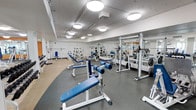 The fitness center is a spacious, single-floor facility designed to support a comprehensive strength training experience. The room is finished with a durable rubber flooring in a neutral gray tone, providing both safety and comfort underfoot. Overhead, circular flush-mount light fixtures are evenly distributed across the white ceiling, ensuring the space is well-lit throughout. A row of large mirrors lines one wall, visually expanding the room while allowing users to monitor their form during exercise. The equipment layout is thoughtfully organized, featuring multiple adjustable weight benches with blue upholstery, squat racks, a dumbbell rack with a full range of free weights, and various cable and resistance machines positioned along the perimeter. The open floor plan allows for easy movement between stations, minimizing congestion during peak use. The clean white walls and bright lighting contribute to an energetic yet functional atmosphere. Overall, the fitness center offers a well-maintained, practical environment suited for a variety of strength and conditioning workouts.