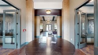 This expansive clubhouse serves as a full-featured community amenity space spread across a generous single-level footprint. The grand entrance greets visitors with soaring ceilings, distinctive textured brick columns, and rich hardwood-style flooring that sets an elegant tone throughout. The open-concept main lounge features dramatic vaulted ceilings with exposed dark beams, a floor-to-ceiling fireplace surround with a mounted flat-screen TV, and bold purple accent walls that create a striking visual contrast against neutral furnishings. Adjacent to the lounge, a well-appointed dining and café area showcases a coffered ceiling detail, pendant lighting, patterned wallcovering, and multiple seating arrangements suited for socializing or casual meetings. A coffee and refreshment bar adds convenience to the space. The fitness center spans two dedicated exercise rooms equipped with cardio machines, stair climbers, and mounted televisions, with large windows providing ample natural light. A stylish game room features an ornate coffered ceiling, foosball table, and high-top seating. Private glass-enclosed offices offer a quiet workspace. Outdoors, a resort-style pool with a central island feature, surrounding palm trees, lounge chairs, and manicured turf areas completes this exceptional amenity offering.
