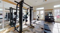Located at 11201 Carmenita Rd in Santa Fe Springs, CA, this fitness center offers a thoughtfully designed workout environment on a single floor. The space features soaring ceilings that lend an open, expansive feel, complemented by an abundance of natural light streaming through large French doors and transom windows along multiple walls. Ceiling fans provide comfortable air circulation throughout the facility.

The gym is comprehensively outfitted with a wide variety of equipment to support diverse fitness routines. Strength training options include a cable machine, free weights, a dumbbell rack, and a dedicated weight bench. Cardio equipment includes treadmills, elliptical trainers, a stair climber, and a rowing machine. A well-stocked rack of medicine balls and stability balls adds functional training capability to the space.

Full-length mirrors line portions of the walls, amplifying the sense of space and allowing users to monitor their form during workouts. The floor is covered in a distinctive geometric-patterned carpet in neutral tones of gray, green, and beige, adding visual interest to the space. A small refreshment counter with a wine cooler and built-in cabinetry is finished with dark tile accents, providing a convenient amenity. The overall ambiance is clean, bright, and welcoming, making it a well-rounded facility for a full-body workout.