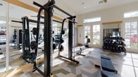 Located at 11201 Carmenita Rd in Santa Fe Springs, CA, this fitness center offers a thoughtfully designed workout environment on a single floor. The space features soaring ceilings that lend an open, expansive feel, complemented by an abundance of natural light streaming through large French doors and transom windows along multiple walls. Ceiling fans provide comfortable air circulation throughout the facility.

The gym is comprehensively outfitted with a wide variety of equipment to support diverse fitness routines. Strength training options include a cable machine, free weights, a dumbbell rack, and a dedicated weight bench. Cardio equipment includes treadmills, elliptical trainers, a stair climber, and a rowing machine. A well-stocked rack of medicine balls and stability balls adds functional training capability to the space.

Full-length mirrors line portions of the walls, amplifying the sense of space and allowing users to monitor their form during workouts. The floor is covered in a distinctive geometric-patterned carpet in neutral tones of gray, green, and beige, adding visual interest to the space. A small refreshment counter with a wine cooler and built-in cabinetry is finished with dark tile accents, providing a convenient amenity. The overall ambiance is clean, bright, and welcoming, making it a well-rounded facility for a full-body workout.
