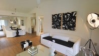 This single-floor space makes a strong visual impression through its vaulted ceilings, which lend a sense of openness and architectural interest to the living area. Warm hardwood flooring runs throughout, providing a grounding contrast to the predominantly white walls and furnishings. The living room is styled in a crisp monochromatic palette, anchored by a white sofa dressed with black accent pillows and complemented by a cowhide ottoman and a glass coffee table. A striking pair of large-scale black and white abstract paintings in natural wood frames serves as a bold focal point on the main wall, adding artistic depth to the otherwise understated décor. Natural light filters into the space, enhancing its airy feel. A hallway visible from the living area leads to what appears to be a bathroom and vanity area, finished in warm wood tones with neutral tile accents, suggesting a cohesive design language throughout. A ceiling fan is suspended from the peak of the vaulted ceiling, adding both function and a casual, relaxed quality. Overall, the space balances minimalist elegance with curated artistic details.