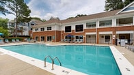 This residential community amenity space is defined by its expansive, curvilinear outdoor swimming pool, which serves as the central focal point of the grounds. The pool features multiple entry points via stainless steel railings and is surrounded by a generous concrete deck furnished with lounge chairs and shaded umbrella stations, offering ample space for relaxation and recreation. The adjoining clubhouse is constructed in a classic brick and white-trim architectural style with a pitched shingle roof, large windows, and an upper-level balcony that overlooks the pool area. A clothes care center and additional resident service facilities are accessible directly from the pool deck. Beyond the pool, the grounds open to thoughtfully landscaped common areas featuring mature trees, manicured shrubs, natural stone drainage features, and winding paved pathways that create a welcoming, park-like atmosphere. A full tennis court is also part of the amenity offering, visible within the secured community grounds. The lush green surroundings, including tall pines and deciduous trees, provide a natural backdrop that enhances the sense of privacy and tranquility throughout the outdoor spaces. The overall ambiance is that of a well-maintained, amenity-rich community designed for active outdoor living.