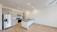 Located in San Tan Valley, AZ, this well-designed one-bedroom, one-bathroom unit offers a clean, contemporary aesthetic with a thoughtful single-floor layout. The open-concept kitchen and living area serves as the heart of the space, featuring light wood-grain cabinetry that extends to the ceiling for generous storage, a white subway tile backsplash, and a full suite of stainless steel appliances including a refrigerator, electric range, and built-in microwave. A central kitchen island with a quartz-style countertop and undermount sink provides additional workspace and doubles as a casual dining area. Light wood-style vinyl flooring flows seamlessly throughout the entire unit, lending warmth and visual continuity. The living area is bathed in natural light through large windows fitted with horizontal blinds, with a glass exterior door offering direct outdoor access. The spacious bedroom features a ceiling fan, a large window with exterior views, and a double-door closet. The bathroom is finished in a matching modern palette, with a wood-grain vanity, integrated sink, and a wall-mounted storage cabinet. Recessed lighting throughout the unit ensures consistent, bright illumination, completing the fresh and functional design of this San Tan Valley residence.