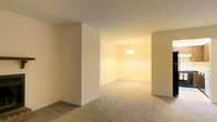 This single-floor space features an open layout connecting multiple living areas through a partially divided floor plan. The interior is finished with wall-to-wall carpeting in a neutral beige tone, providing a consistent and cohesive look throughout. The walls are painted in soft, warm cream tones that complement the overall color palette.

A partial wall divides the main living area from an adjacent room, which is illuminated by a centrally hung pendant light fixture. This pendant light serves as a focal point in the secondary space, casting a warm ambient glow that enhances the inviting atmosphere. The rooms are separated by defined architectural transitions rather than full walls, preserving a sense of openness and flow between spaces.

The ceilings feature a textured finish throughout, and the space benefits from warm artificial lighting that highlights the neutral color scheme. Chair rail-style molding is visible along the walls of the rear room, adding a subtle decorative architectural detail.

Overall, the space presents a clean, understated interior with practical finishes and a warm, approachable ambiance suited to everyday living.
