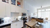 This single-floor space showcases a thoughtfully designed open-concept layout that seamlessly connects the kitchen, dining, and living areas. The kitchen is outfitted with white cabinetry extending toward impressively tall ceilings, creating a sense of vertical spaciousness. A classic white subway tile backsplash complements the granite countertops, while the stainless steel appliances — including a range and built-in microwave — provide a sleek, contemporary aesthetic. A double-basin sink with a modern faucet adds functional elegance to the space.

Adjacent to the kitchen, a rustic wooden dining table with seating anchors the dining area, offering a warm contrast to the clean, modern finishes throughout. The living area beyond benefits from a large window that draws in generous natural light, enhancing the bright, airy atmosphere. Decorative wall art and thoughtful accessory placements add personality to the space. The consistently white walls and high ceilings throughout contribute to an open, uncluttered ambiance that feels both welcoming and practical for everyday living.