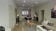 This single-floor space showcases a clean, contemporary design centered around an open-plan layout that connects the living, dining, and kitchen areas in one cohesive environment. Polished concrete floors run throughout, lending an industrial-modern aesthetic that is both durable and visually striking. Recessed ceiling lights are positioned throughout the space, providing consistent, well-distributed illumination across all zones.

The kitchen features white cabinetry and a bright window that allows natural light to fill the area, complementing the neutral white walls that define the overall palette. Adjacent to the kitchen, the dining area accommodates a round white table surrounded by modern Eames-style chairs, creating a functional yet stylish gathering space. Black shelving units flank the dining zone, offering display and storage while reinforcing the contrast between dark accents and the lighter surroundings.

The living area flows naturally from the dining space, anchored by a light area rug and a low white coffee table. A sculptural wooden piece leans against the wall near the entry, adding an artistic, organic element to the otherwise minimalist interior. Framed artwork on the walls contributes to the curated, gallery-like atmosphere. The open layout and cohesive design make this a versatile and well-appointed single-level space.