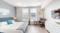 This single-floor unit offers a thoughtfully designed layout that balances comfort and functionality across its key spaces. The bedroom serves as the central living area, featuring light wood-style flooring, soft neutral-toned walls, and dual windows fitted with horizontal blinds that allow ample natural light to filter in. A tufted blue upholstered bed frame anchors the room, complemented by matching glass and metal nightstands with table lamps. A cozy seating nook includes a brown wingback chair paired with an arc floor lamp and a small side table, while a white dresser provides additional storage. An accent wall in muted blue-grey adds depth to the space.

The compact kitchenette is efficiently organized with two-tone cabinetry in light blue and grey, open shelving, a flat-top cooktop, an under-counter microwave, a full-size refrigerator, and a single-basin sink — providing all essential amenities within a streamlined footprint.

The bathroom is clean and modern, featuring a white floating vanity with a rectangular undermount sink, a large mirror with overhead lighting, a shower with curtain, and wood-style flooring. A colorful abstract artwork adds a subtle decorative touch. Overall, the space maintains a cohesive, contemporary aesthetic throughout.