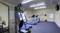 This single-story fitness room offers a practical and functional space designed to support a range of workout routines. The room is outfitted with multiple treadmills and cardio machines arranged in a row, providing ample opportunity for cardiovascular exercise. A multi-station strength training machine anchors one side of the room, offering users a variety of resistance exercises in a compact footprint. The flooring consists of durable blue carpet, well-suited for a fitness environment. Overhead fluorescent lighting ensures the space is brightly and evenly illuminated throughout. A window along one wall allows for a degree of natural light, contributing to a more open and comfortable atmosphere. Informational posters and a bulletin board are mounted on the walls, offering exercise guidance and community notices. A ceiling fan aids in air circulation, enhancing comfort during workouts. The layout is efficient and uncluttered, making it easy to move between equipment. Overall, the space delivers a straightforward and accessible fitness facility with the essential equipment needed for a balanced exercise routine.