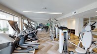 This single-floor fitness center offers a thoughtfully designed workout environment that combines functionality with a clean, contemporary aesthetic. The space features a bright, open layout with linear LED lighting running across the white ceiling, creating an energizing and well-lit atmosphere throughout.

The gym is generously outfitted with a wide array of cardio equipment, including a row of elliptical trainers and additional aerobic machines positioned along the perimeter. Complementing the cardio section, a robust selection of Hoist-brand strength training machines is arranged across the floor, providing users with a comprehensive full-body workout experience. A dedicated free weights and storage area with wooden shelving units adds further versatility to the space.

Mounted flat-screen televisions are positioned throughout the facility, allowing members to stay entertained during their workouts. The herringbone-patterned wood-look flooring adds a refined, upscale touch that sets this gym apart from a standard fitness space. Large windows along one wall invite natural light and contribute to an open, airy feel. The neutral color palette of whites and warm wood tones creates a clean and motivating environment well-suited for daily exercise routines.