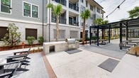 Located at 350 Henslow Dr in Charleston, South Carolina, this thoughtfully designed outdoor amenity courtyard offers residents a refined and functional gathering space surrounded by the natural beauty of the Lowcountry. The space is anchored by multiple stainless steel outdoor grilling stations built into handsome brick-clad counters, providing ample cooking space for entertaining. A striking dark-framed pergola with slatted overhead beams creates a shaded seating area outfitted with a large dining table and benches, ideal for al fresco meals. String lights are strung overhead, adding warmth and ambiance for evening gatherings. The paved courtyard features a mix of herringbone-patterned pavers and concrete surfaces, bordered by carefully maintained landscaping with mature live oaks, tall palms, and fresh mulched planting beds. Adirondack seating areas are positioned throughout the open courtyard, encouraging relaxed outdoor enjoyment. The space is framed by the surrounding multi-story residential building, whose gray-toned facades, white brick accents, and private balconies overlook the courtyard. The overall atmosphere is clean, modern, and welcoming, blending Charleston's architectural character with contemporary outdoor living amenities.