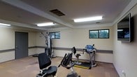 This single-floor fitness facility offers a practical and functional workout environment designed to accommodate a range of exercise needs. The room is outfitted with several pieces of equipment, including a multi-station strength training machine, a rowing machine, and a stationary recumbent bike, providing options for both cardiovascular and resistance training. The flooring features a combination of carpet and hard surface areas, offering comfortable footing across different workout zones. Bright fluorescent lighting fixtures are mounted across the ceiling, ensuring the space is evenly and thoroughly illuminated. A series of horizontally oriented windows line the upper portion of the walls, allowing natural light to filter in and providing views of the surrounding outdoor greenery. The walls are finished in a neutral tone with a dark accent border running along the base, lending the room a clean and utilitarian aesthetic. Ventilation is supported by ceiling-mounted air ducts, contributing to a comfortable exercise environment. The overall layout is open and uncluttered, allowing users to move freely between equipment and making the space accessible and easy to navigate.