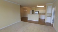 This single-floor residence showcases an open-concept layout that seamlessly connects the living, dining, and kitchen areas. The living area is finished with plush, light-toned carpeting, creating a comfortable and inviting atmosphere, while the kitchen and dining zones transition to warm hardwood-style flooring that adds a refined contrast.

The kitchen is well-appointed with white cabinetry offering ample storage, a stainless steel refrigerator, and a central island that serves as both a functional workspace and a natural divider between the cooking and living spaces. Recessed lighting throughout the kitchen and dining area ensures bright, even illumination, while a classic chandelier above the dining space adds an elegant focal point.

Crown molding runs along the upper edges of the walls, lending the space a polished, traditional character. The neutral warm-beige wall tones throughout create a cohesive and welcoming environment. The open floor plan allows natural sightlines across all zones, making the space feel expansive and functional for everyday living and entertaining.