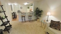 This single-floor residence offers an open-concept layout that connects the living, dining, and kitchen areas into one cohesive and functional space. The living area features plush wall-to-wall carpeting in a warm neutral tone, creating a comfortable and inviting atmosphere. A glass-top side table and soft lamp add refined touches to the space.

The kitchen is positioned at the heart of the home, highlighted by a raised breakfast bar with elegant wrought-iron swivel bar stools with cushioned seats — ideal for casual dining or entertaining. White cabinetry with upper shelving extends to the ceiling, offering ample storage, while decorative greenery adds a lively accent above the cabinets. The kitchen connects seamlessly to a separate dining area, visible in the background, which features additional seating and hardwood-style flooring.

The overall color palette is light and neutral, with soft white walls and warm-toned finishes throughout, contributing to a bright and airy ambiance. Flush-mounted ceiling lights provide consistent illumination across the space. A large decorative indoor tree adds a natural, organic element to the living area, enhancing the welcoming character of the home. The layout is thoughtfully designed to maximize both flow and functionality.