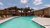 Located at 8787 Hammerly Blvd in Houston, Texas, this outdoor amenity space centers around a generously sized swimming pool with crystal-clear turquoise water, marked at a depth of 8 feet. The pool is framed by a wide concrete deck that accommodates multiple rows of modern sling-style lounge chairs, providing ample space for sunbathing and relaxation. Overhead, bold blue shade canopies supported by white steel frames offer welcome relief from the Texas sun, creating comfortable shaded lounging areas directly adjacent to the pool. Tall, mature palm trees are strategically planted throughout the area, reinforcing a resort-like tropical atmosphere. The surrounding grounds are well-maintained, featuring neatly landscaped green lawns and iron perimeter fencing that defines the pool area. The backdrop consists of a vibrant multi-building residential community, with structures finished in a lively palette of teal, yellow, orange, and red accents that contribute to the space's energetic and welcoming character. Exterior staircases and balconies are visible on the surrounding two-story buildings, adding architectural depth to the setting. The entire outdoor environment benefits from abundant natural light and open sky views, making it a dynamic and inviting communal gathering space suited to the warm Houston climate.