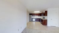This single-floor space offers a clean, open-concept layout that seamlessly connects the living area to the kitchen, creating a sense of openness and ease of movement throughout. The kitchen is a standout feature, outfitted with rich dark wood cabinetry that provides generous storage space. Stainless steel appliances — including a refrigerator, dishwasher, microwave, and range — lend a modern, polished look to the space. A double-basin sink is positioned beneath overhead cabinetry, and light countertops provide a bright contrast to the deep-toned cabinets. The kitchen flooring transitions to a light-toned surface, while the adjacent living area is finished with neutral carpeting, offering a comfortable underfoot feel. The walls throughout are painted in a crisp, neutral white, contributing to a bright and airy ambiance. Overhead fluorescent lighting in the kitchen ensures the workspace is well-lit. The overall design is functional and straightforward, with a neutral palette that provides a versatile backdrop for a variety of interior styles. The open flow between the kitchen and living space makes this a practical and welcoming environment.