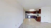 This single-floor space offers a clean, open-concept layout that seamlessly connects the living area to the kitchen, creating a sense of openness and ease of movement throughout. The kitchen is a standout feature, outfitted with rich dark wood cabinetry that provides generous storage space. Stainless steel appliances — including a refrigerator, dishwasher, microwave, and range — lend a modern, polished look to the space. A double-basin sink is positioned beneath overhead cabinetry, and light countertops provide a bright contrast to the deep-toned cabinets. The kitchen flooring transitions to a light-toned surface, while the adjacent living area is finished with neutral carpeting, offering a comfortable underfoot feel. The walls throughout are painted in a crisp, neutral white, contributing to a bright and airy ambiance. Overhead fluorescent lighting in the kitchen ensures the workspace is well-lit. The overall design is functional and straightforward, with a neutral palette that provides a versatile backdrop for a variety of interior styles. The open flow between the kitchen and living space makes this a practical and welcoming environment.