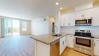 Located at 310-340 Palm Ave in National City, California, this single-story unit offers a clean, contemporary design throughout. The open-concept layout connects the kitchen and living area seamlessly, creating a bright and functional living space. Light wood-tone flooring runs throughout, complementing the neutral white walls and recessed lighting overhead.

The kitchen is well-appointed with white shaker-style cabinetry featuring matte black hardware, a white subway tile backsplash, and quartz countertops. A peninsula island with an undermount sink and a high-arc pull-down faucet provides additional prep space and casual seating potential. Stainless steel appliances include a refrigerator, electric range, built-in microwave, and dishwasher. The living area is equipped with a wall-mounted mini-split system for climate control and connects to an outdoor area through a sliding glass door with a screen.

The bedroom features generous wall space, a window with views of the surrounding neighborhood, and direct access to a walk-in closet and an en-suite bathroom. The bathrooms include white vanities with integrated countertops, mirror medicine cabinets, and a tub/shower combo, all finished with consistent, clean-lined fixtures throughout.