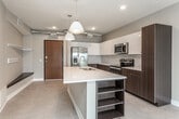 This thoughtfully designed single-floor unit offers a contemporary open-concept layout that seamlessly connects the kitchen and living areas. The kitchen is a standout feature, showcasing a large center island with a built-in sink and crisp white countertops, complemented by a striking two-tone cabinet design — dark espresso lower cabinets and bright white upper cabinets. A geometric patterned tile backsplash adds a modern touch, while a full suite of stainless steel appliances, including a French door refrigerator with ice maker, electric range, and microwave, rounds out the space. Pendant lighting above the island adds warmth and definition to the area. Exposed ductwork along the ceiling lends an industrial character throughout the open living and kitchen zones. The living area is bright and open, featuring a ceiling fan, neutral flooring, and sliding glass doors that lead to a private balcony with open urban views. A separate bedroom offers plush patterned carpeting and a large picture window framing an expansive cityscape. The bathroom and utility corridor include an in-unit washer and dryer along with ample closet storage. Overall, the unit presents a clean, modern aesthetic with functional, well-appointed spaces throughout.